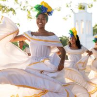 Dancers in white dresses and colourful headwraps perform in front of a white church