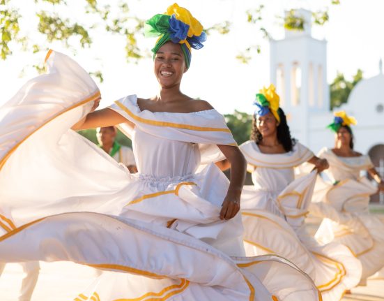 Dancers in white dresses and colourful headwraps perform in front of a white church