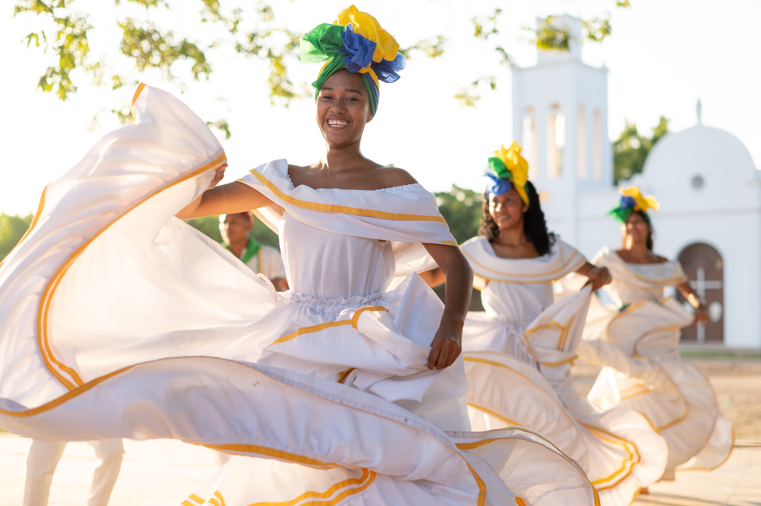 Dancers in white dresses and colourful headwraps perform in front of a white church